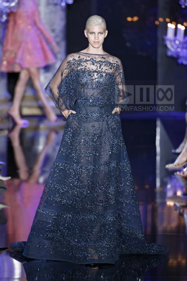 A model on a runway wearing a floor-length navy blue sheer gown with heavy beadwork and a thin waist belt.