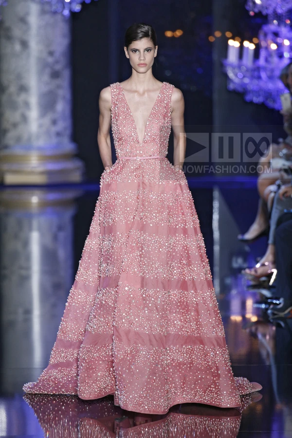 A model on a runway wearing a pink sleeveless gown with a deep V-neckline, fully embellished with pearls and crystals.