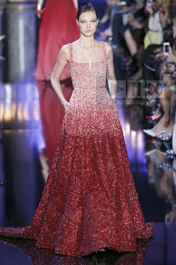A model walks the runway in a floor-length, sleeveless gown with an ombré effect transitioning from light pink to deep red.