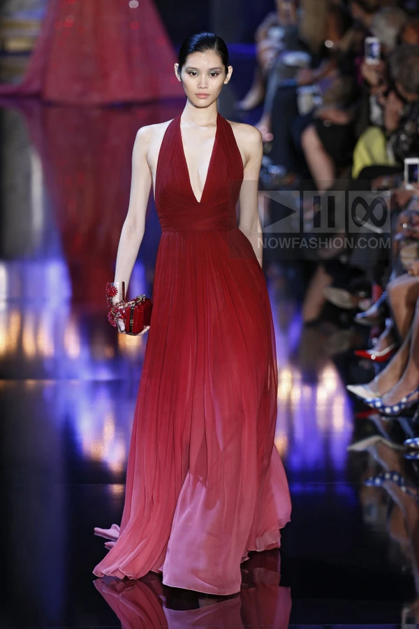 A model walks the runway in a floor-length, deep red, pleated halter-neck gown with a matching clutch.