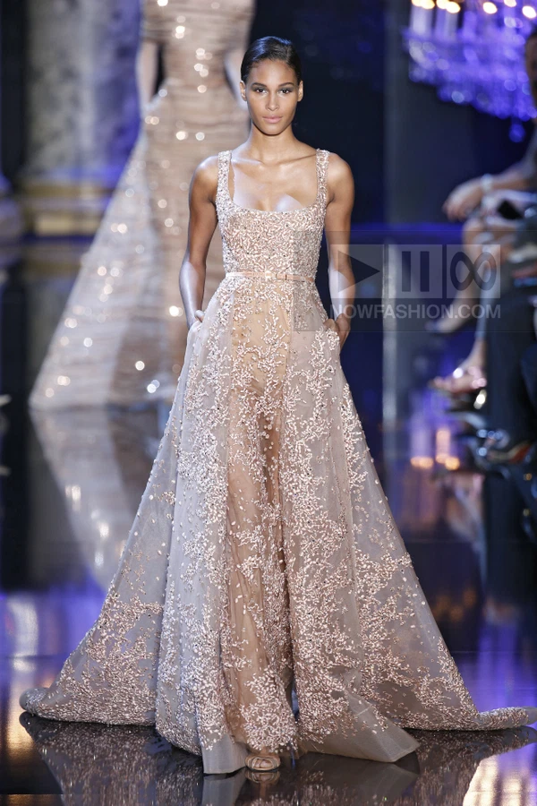 A model walks the runway in a floor-length, blush-colored gown with intricate beadwork and a sheer skirt overlay.