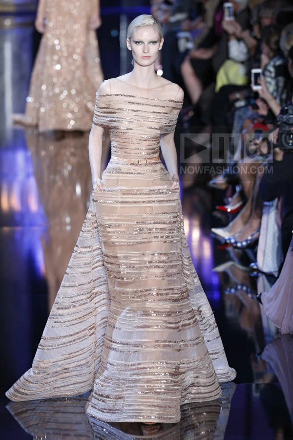 A model on a runway wearing a floor-length, off-the-shoulder gown made of sheer fabric with horizontal sequin stripes.
