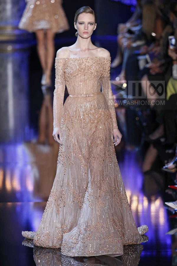 A model walks the runway in a floor-length, off-the-shoulder, nude-colored gown heavily embellished with pearls and beads.