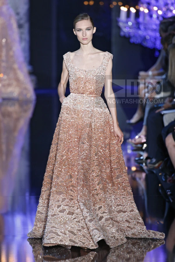 A model walks the runway in a floor-length, sequined, blush-colored gown with a scoop neckline and a thin waist belt.