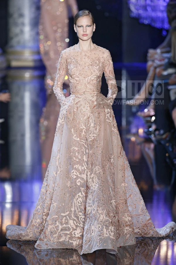 A model walks the runway in a floor-length, long-sleeved gown made of sheer, nude-colored tulle with intricate embroidery.