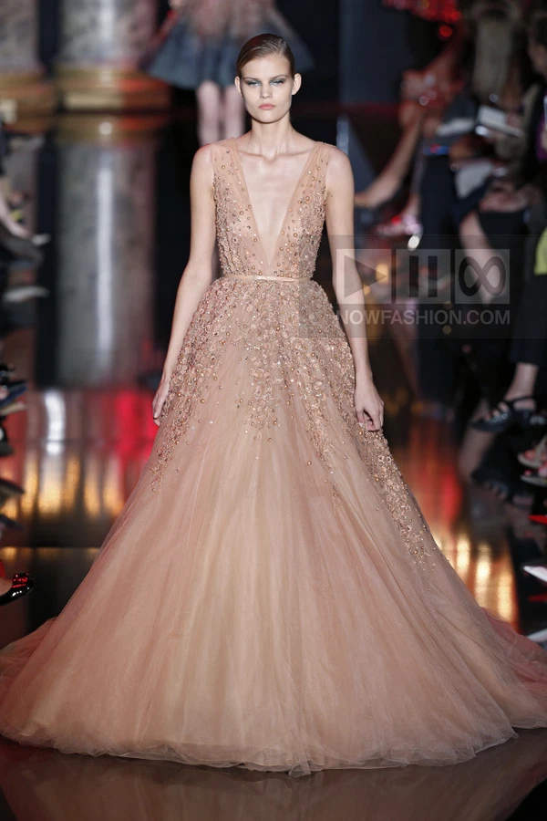 A model wears a floor-length, nude-colored tulle gown with a plunging neckline and dense beadwork on the bodice and skirt.