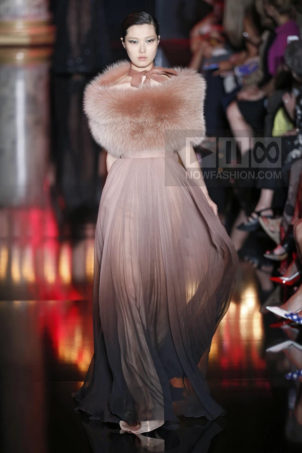 A model walks the runway in a floor-length, pleated, ombre-toned gown with a voluminous fur stole around the shoulders.