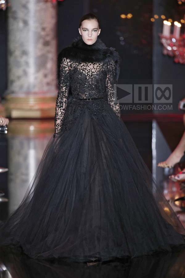 Model on runway wearing a black floor-length gown with long sleeves, intricate beadwork, and a dark fur collar.