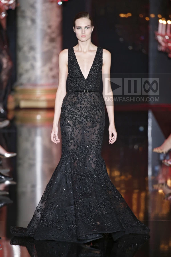A model walks the runway in a black sleeveless floor-length gown with a deep V-neckline and a cinched waist.