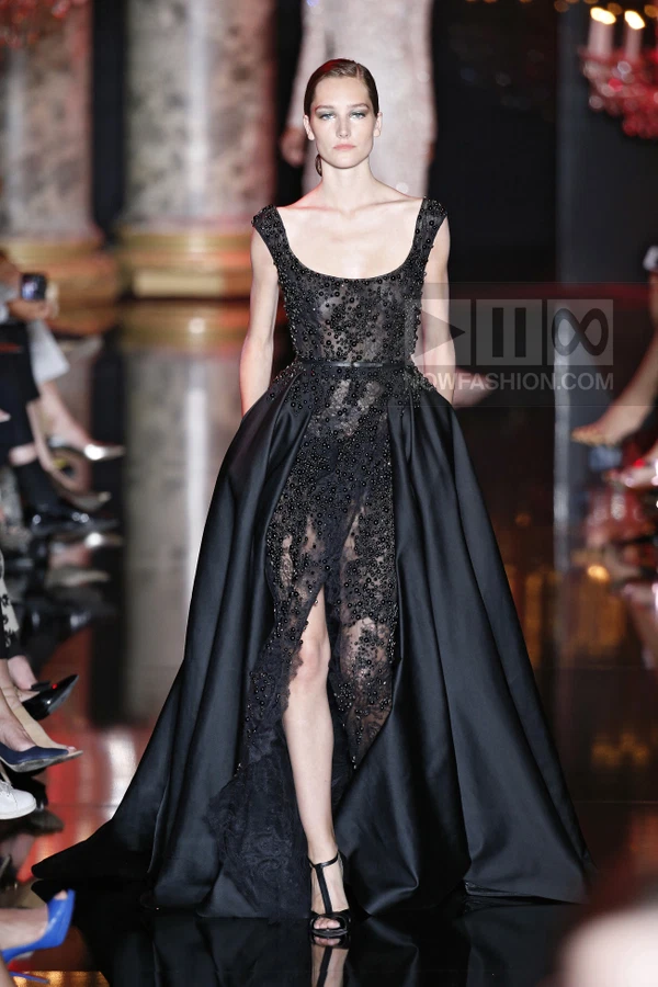 A model walks the runway in a black floor-length gown featuring a beaded lace bodice and a voluminous satin overskirt.
