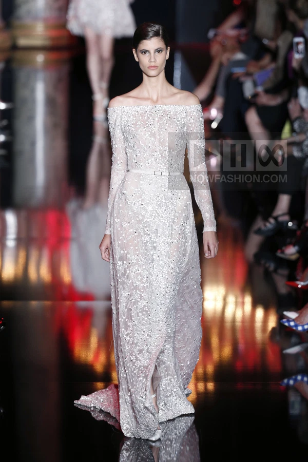 Model wearing a floor-length off-the-shoulder gown with intricate floral embroidery and a thin waist belt on a runway.