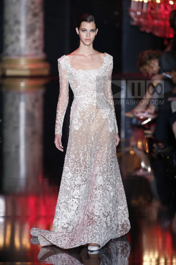 A model walks the runway in a floor-length white lace gown with long sleeves, a square neckline, and a thin waist belt.