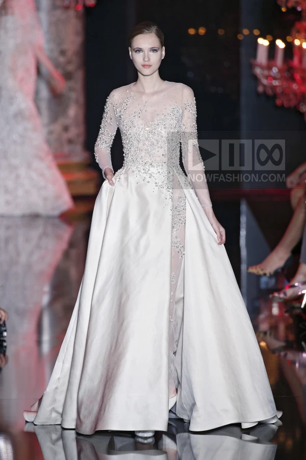 Model on runway wearing a floor-length white gown with sheer bodice, long sleeves, and heavy pearl and crystal embellishment.
