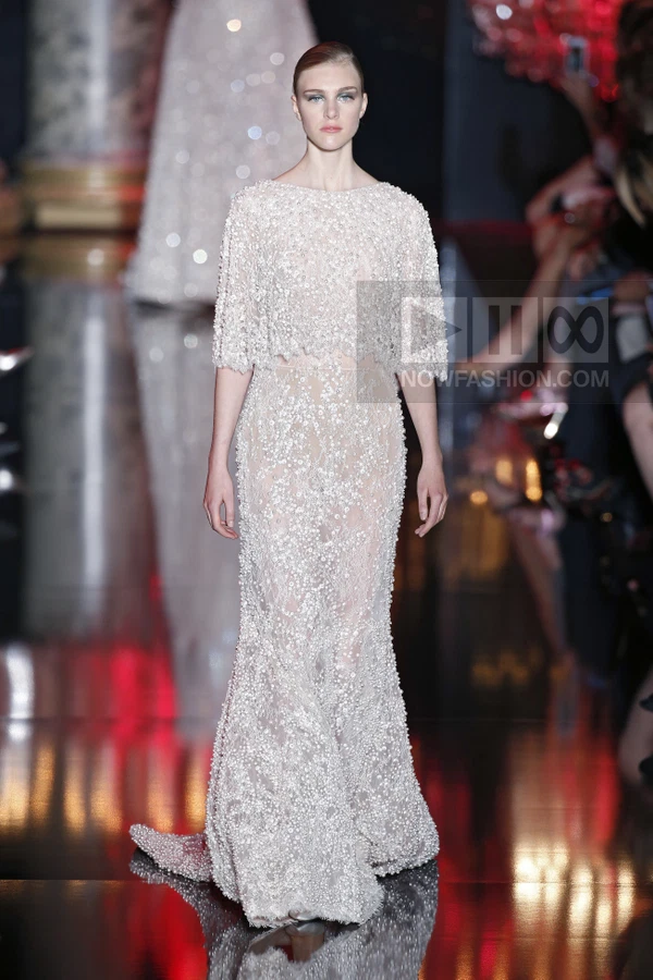 Model on runway wearing a floor-length, beaded white gown with a cropped overlay top and sheer skirt section.