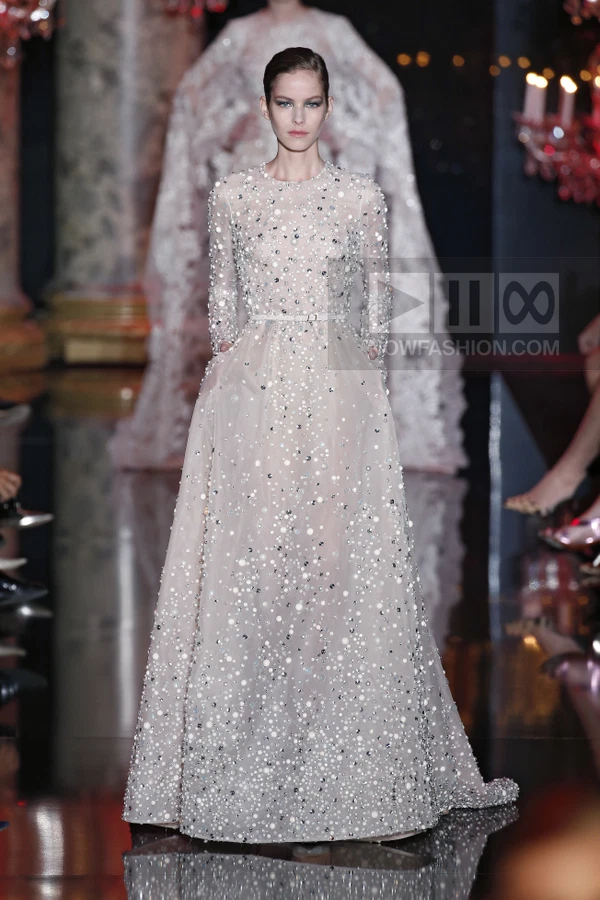 A model walks the runway in a floor-length, long-sleeved, pale pink tulle gown heavily embellished with silver sequins.