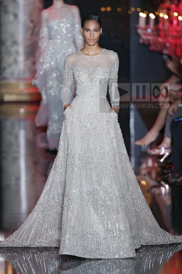 A model walks the runway in a floor-length, light gray, long-sleeved gown heavily embellished with beads and sequins.
