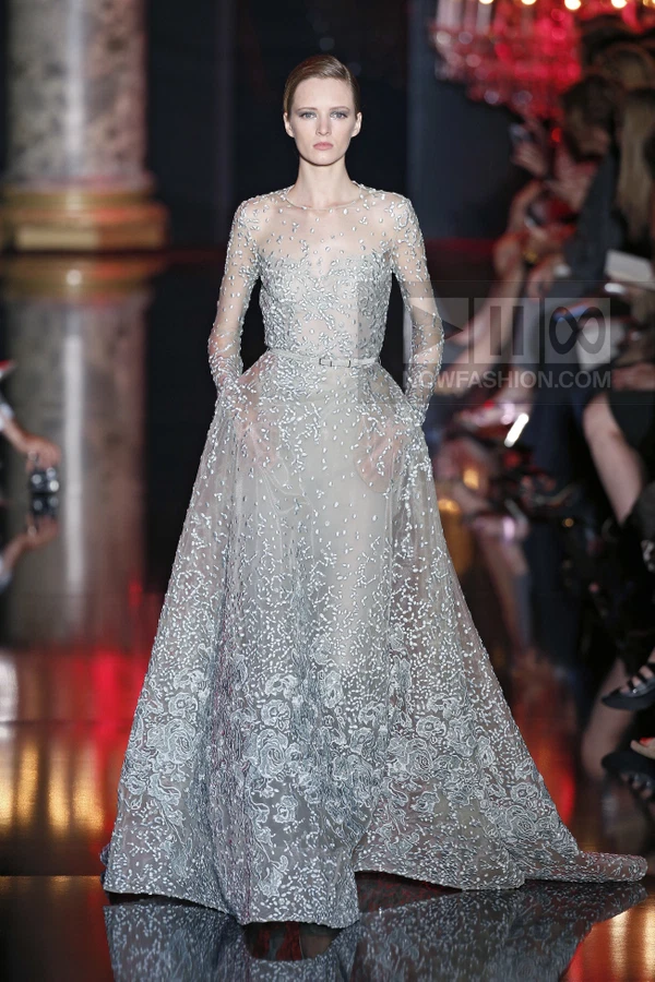 A model walks the runway in a floor-length, light gray gown with intricate silver embroidery and a thin waist belt.
