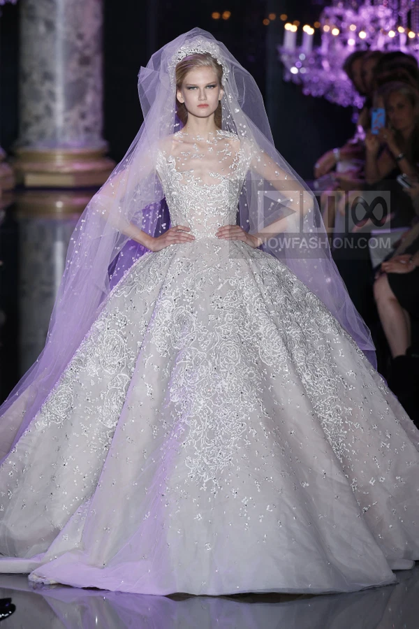 A model wearing a voluminous white ball gown with intricate silver embroidery and a matching veil on a runway.