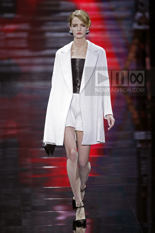 Model walking in a white textured coat, black strapless top, and white shorts on a dark runway with red lighting.