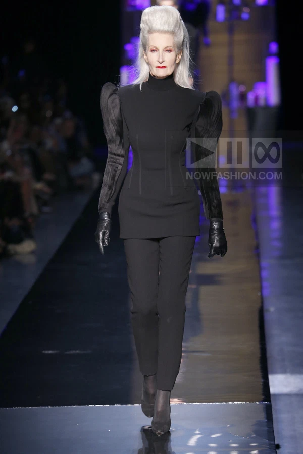Model on runway wearing black long-sleeve top with puffed shoulders, black trousers, and black leather gloves.