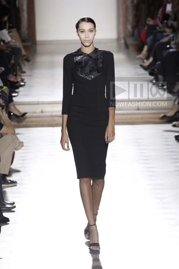 A model walks the runway in a black knee-length sheath dress with a dark floral-patterned chest panel.