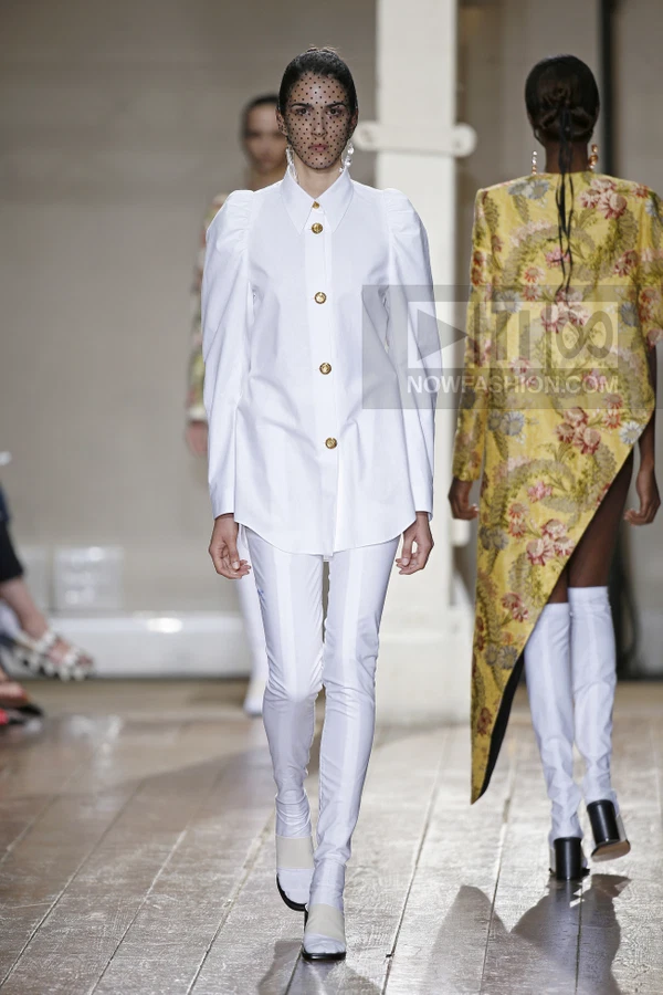 A model walks the runway in a white long-sleeve shirt with puffed shoulders, white trousers, and a polka-dot face veil.