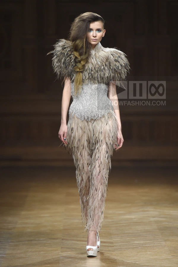 Model wearing a feathered bodice and trousers with a metallic silver corset on a runway.