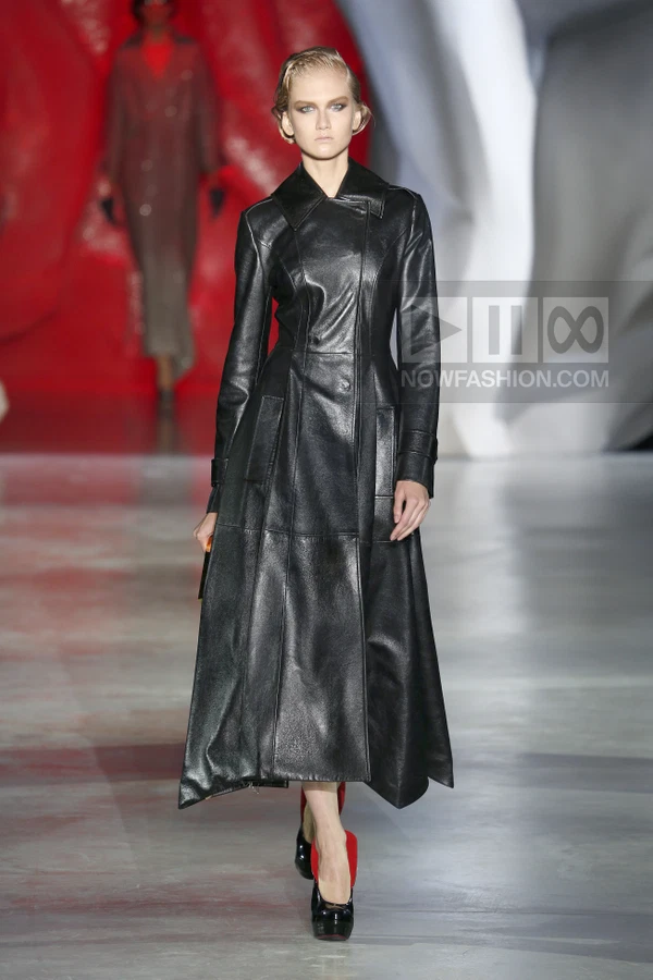 A model walks the runway in a black leather trench coat with a fitted bodice and flared skirt, paired with black pumps.