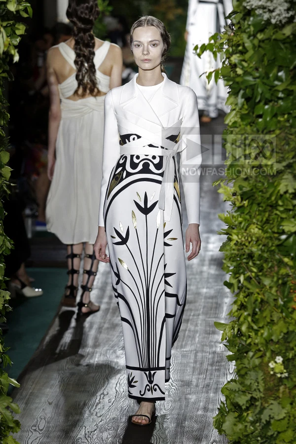 Model walking in a white floor-length dress with black graphic floral print and gold leaf accents, wearing flat sandals.