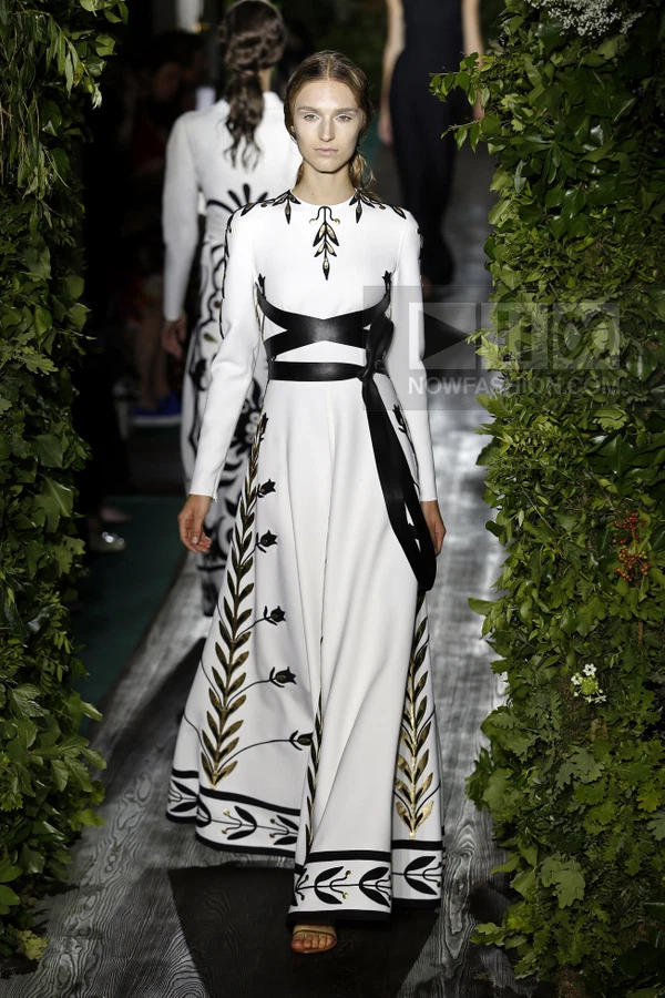 Model walking in a white floor-length gown with black leather waist ties and gold leaf embroidery on a runway.