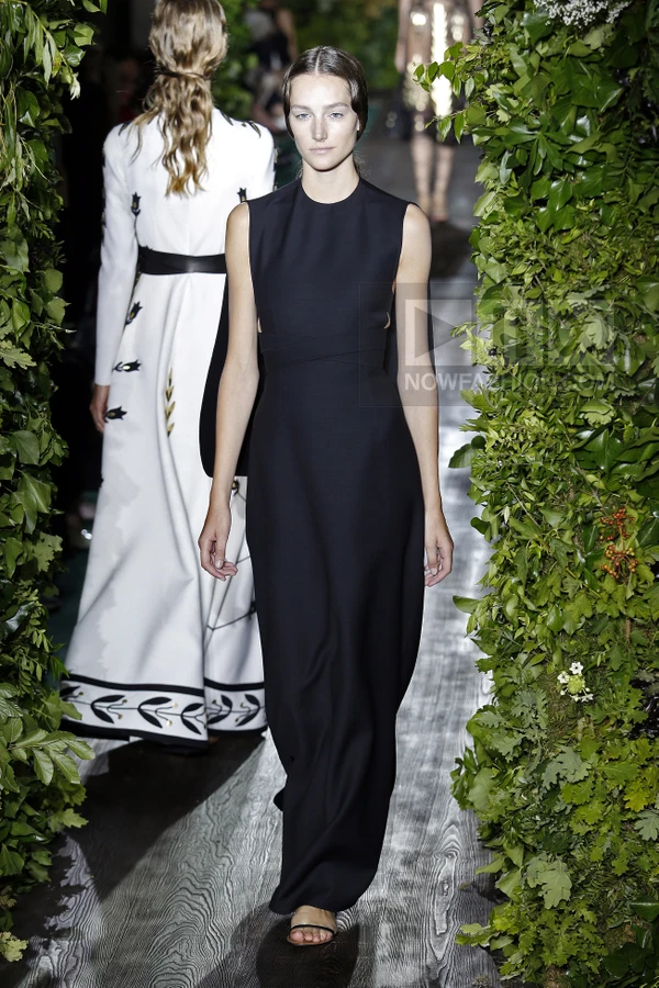 A model walks a runway in a floor-length black sleeveless gown with a cinched waist, flanked by lush green foliage.