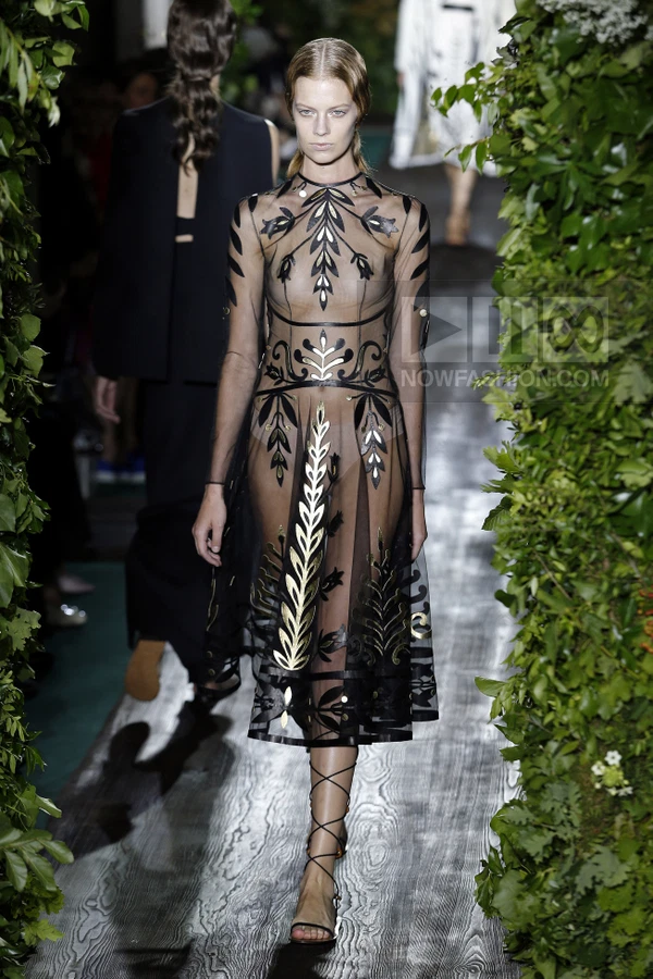 Model walking in a sheer black midi dress with gold and black leaf-patterned appliqués, worn with lace-up sandals.