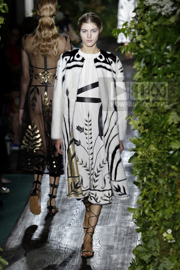 Model walking in a white fur coat with black abstract patterns over a white dress with black and gold botanical embroidery.