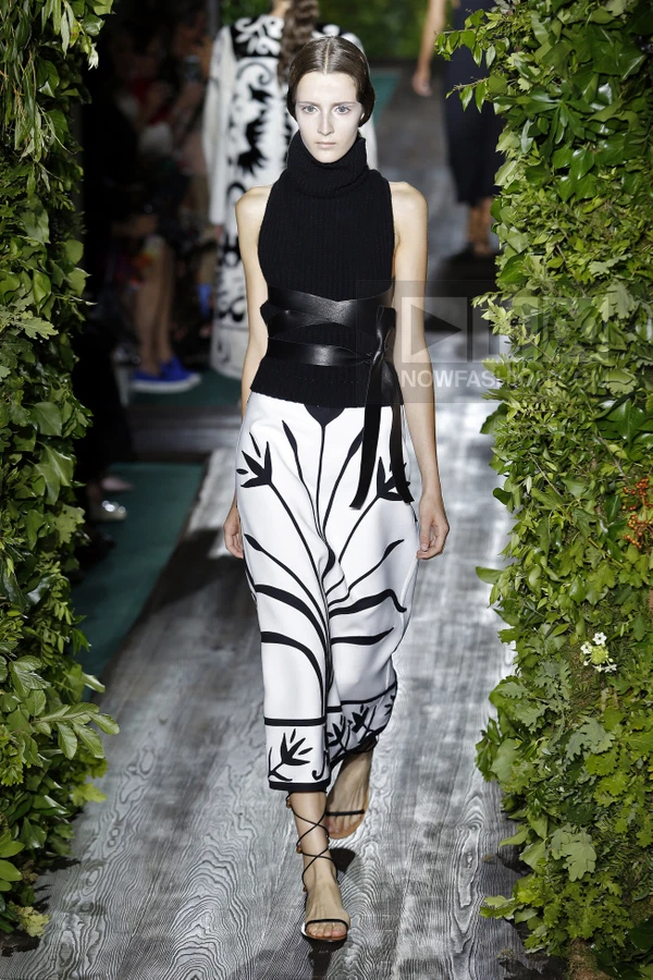 A person walks on a runway wearing a black ribbed turtleneck top, a wide black leather waist belt, and a white midi skirt.