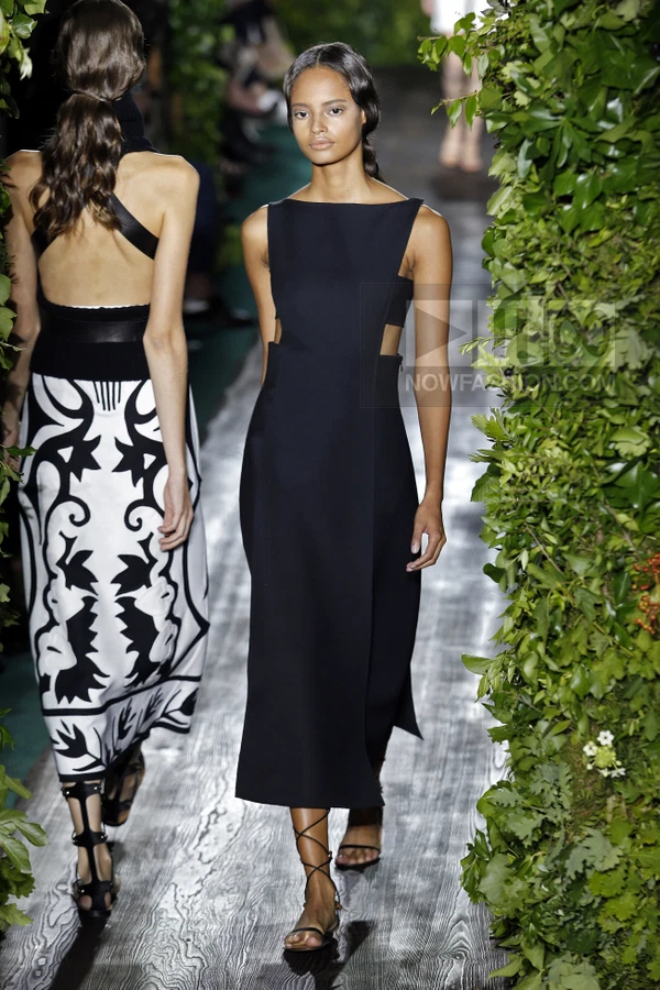 Model walking on a runway wearing a black sleeveless midi dress with side cut-outs and lace-up sandals.