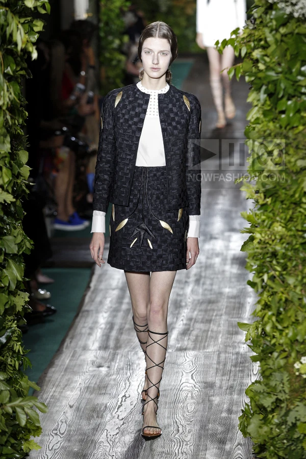 A person walks on a runway wearing a black textured jacket and matching mini skirt with gold leaf-shaped appliqués.