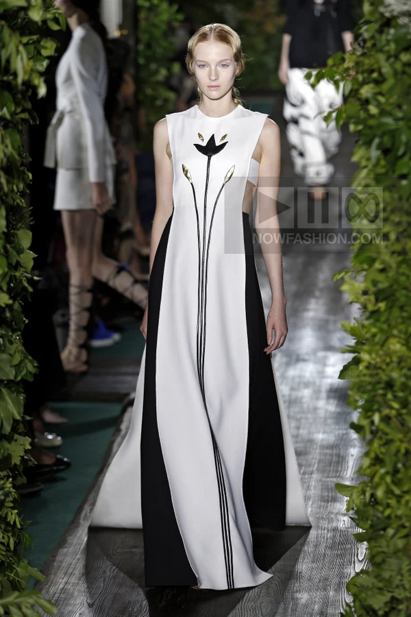 A model walks a runway in a floor-length white and black sleeveless gown with a central floral appliqué.