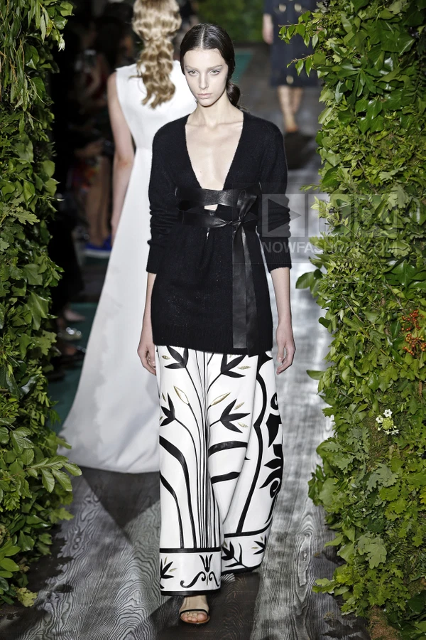 Model walking on a runway wearing a black v-neck sweater with a leather waist tie and wide-leg white trousers with black…