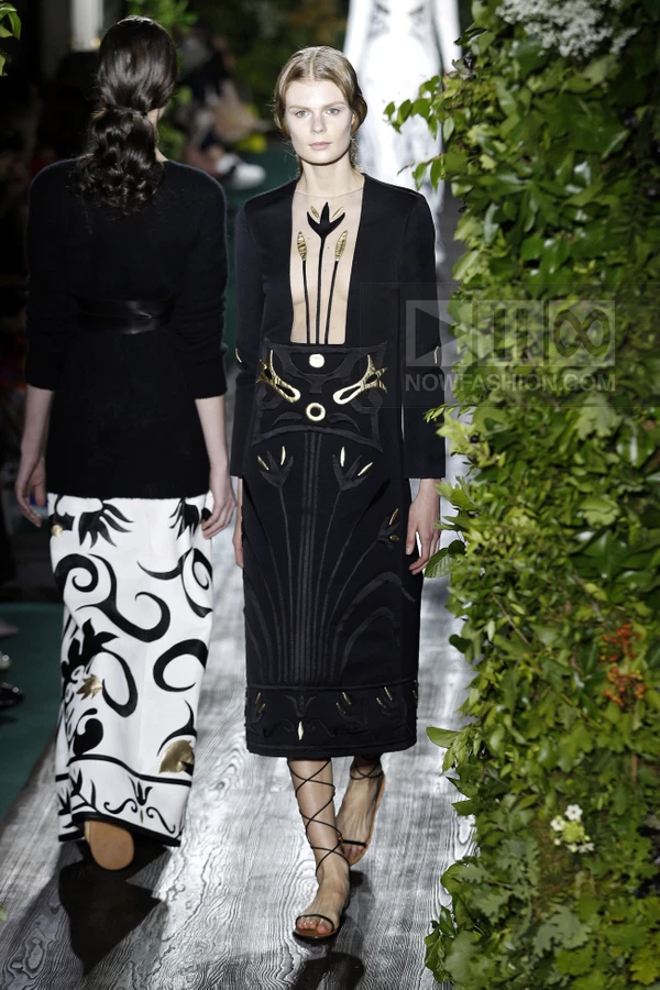 Model walking in a black midi dress with gold floral embroidery and a deep neckline, paired with lace-up sandals.
