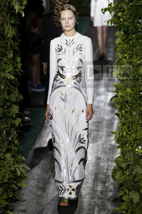 Model walking in a white floor-length dress with silver and gold metallic floral appliqué on a runway.