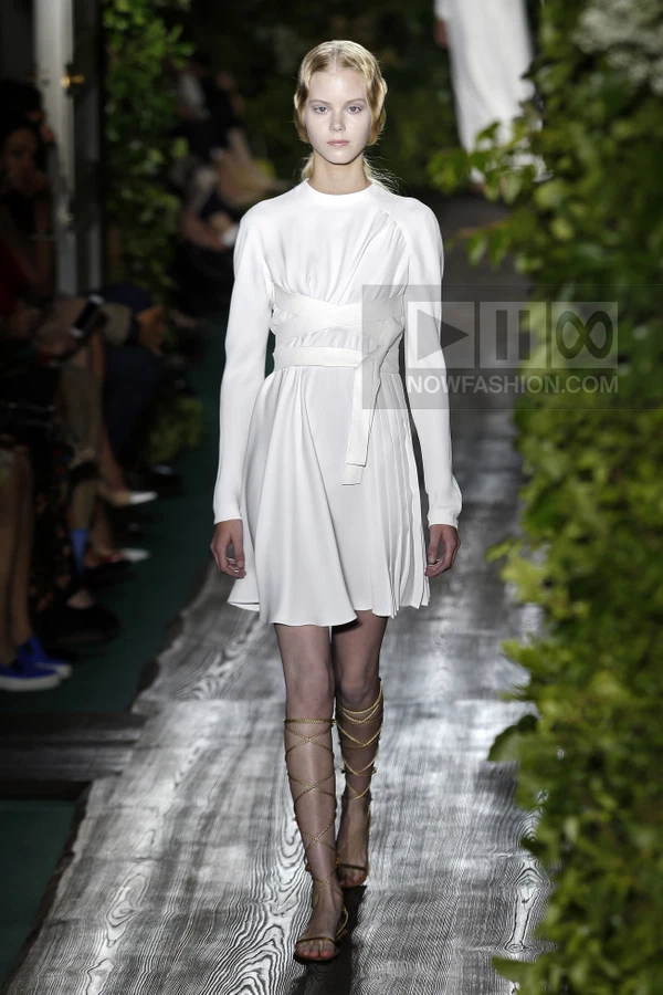 Model wearing a white mini dress with long sleeves and a wrapped waist detail, paired with gold lace-up gladiator sandals.