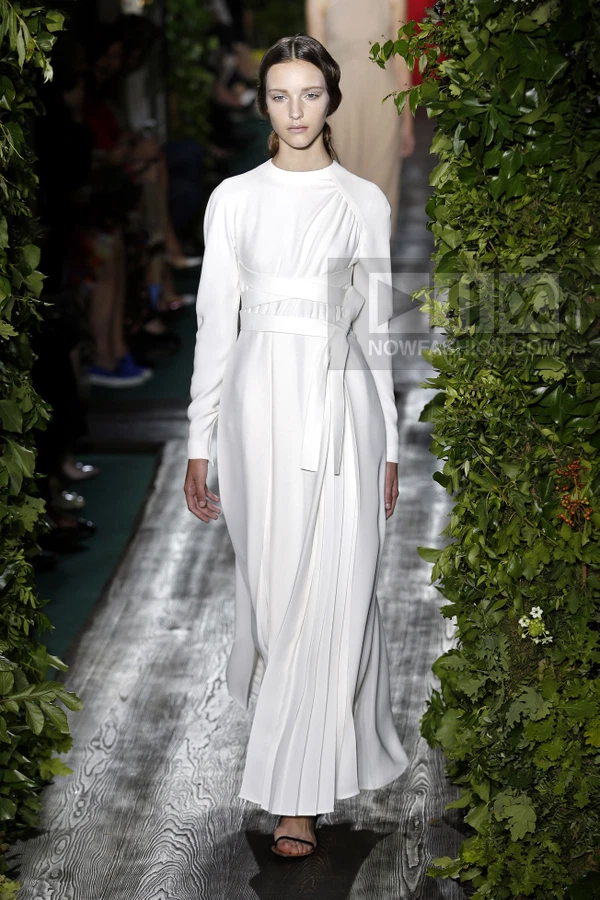 Model in a white long-sleeve floor-length dress with a wrapped bodice and pleated skirt walking on a runway.