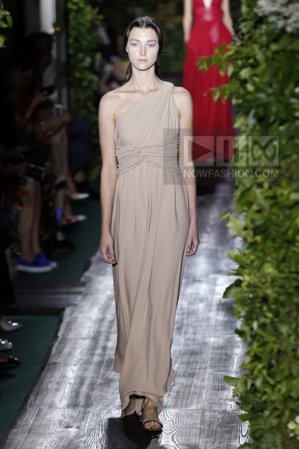 A model walks the runway in a floor-length, one-shoulder beige gown with a draped, knotted bodice.