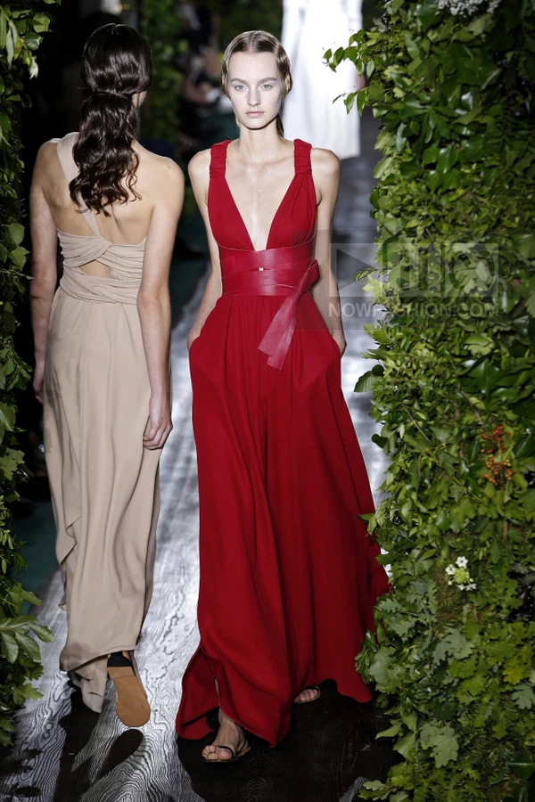 Two models walking on a runway, one in a red sleeveless gown with a leather waist belt, the other in a beige gown.