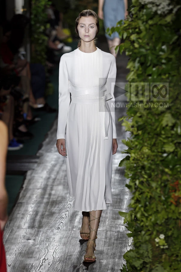 A model walks the runway in a white long-sleeved midi dress with vertical pleats and a matching leather waist tie.