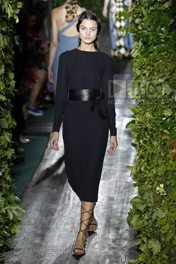 Model walking on a runway wearing a black long-sleeve midi dress with a wide leather waist belt and lace-up sandals.