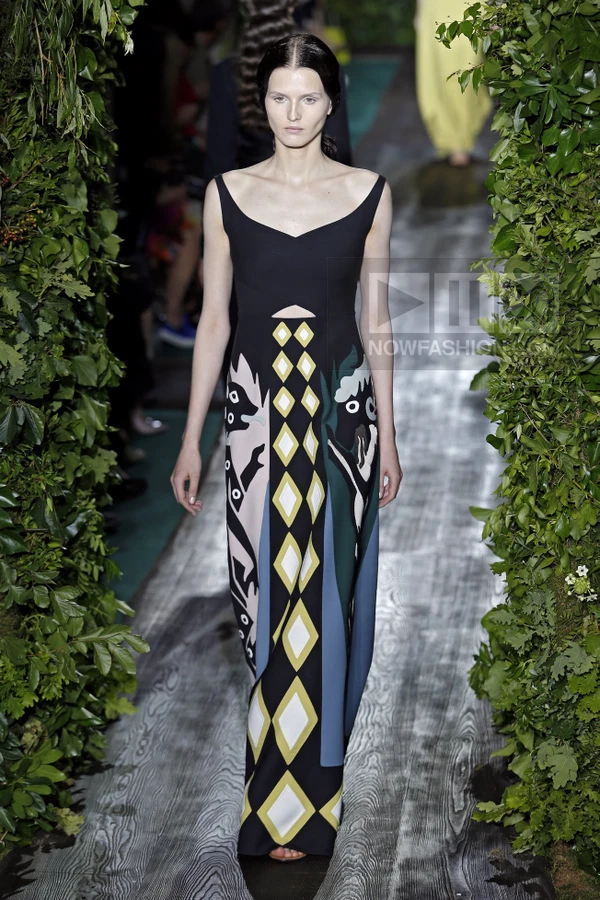 A model walks a runway wearing a black floor-length dress with a geometric diamond pattern and stylized animal motifs.