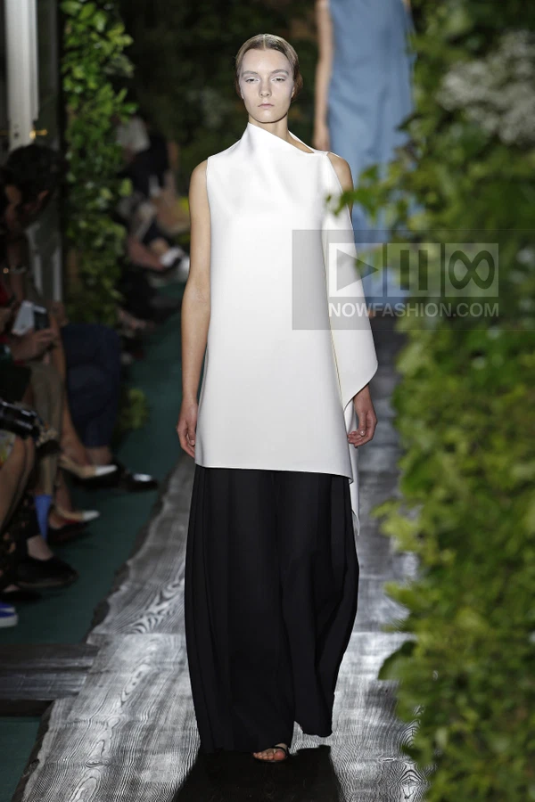 Model walking in a white sleeveless tunic over black wide-leg trousers on a runway with greenery.
