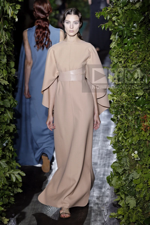 Model in a floor-length beige wool dress with cape sleeves and a wide leather waist belt walking on a runway.