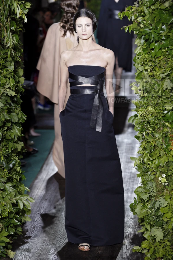 Model wearing a black strapless gown with a leather waist tie and sandals, walking on a runway flanked by greenery.